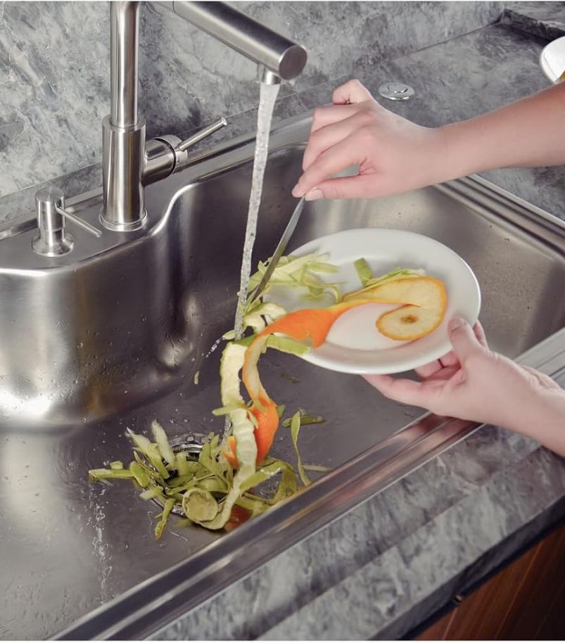 o-que-e-um-triturador-de-alimentos O triturador de alimentos é um eletrodoméstico moderno que elimina resíduos na hora, transformando-os em minúsculas partículas, pessoa despejando restos de alimentos (cascas de laranja e legumes) de um prato diretamente no ralo de uma pia de aço inoxidável com água corrente, ilustrando o funcionamento do triturador de alimentos.