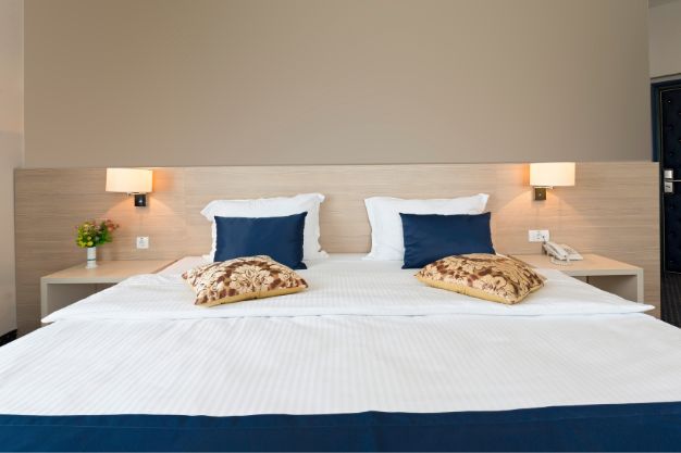 Cama de casal com um amplo colchão queen size, lençóis brancos e travesseiros azuis em um quarto de hotel bem decorado, mostrando as dimensões generosas do colchão queen.