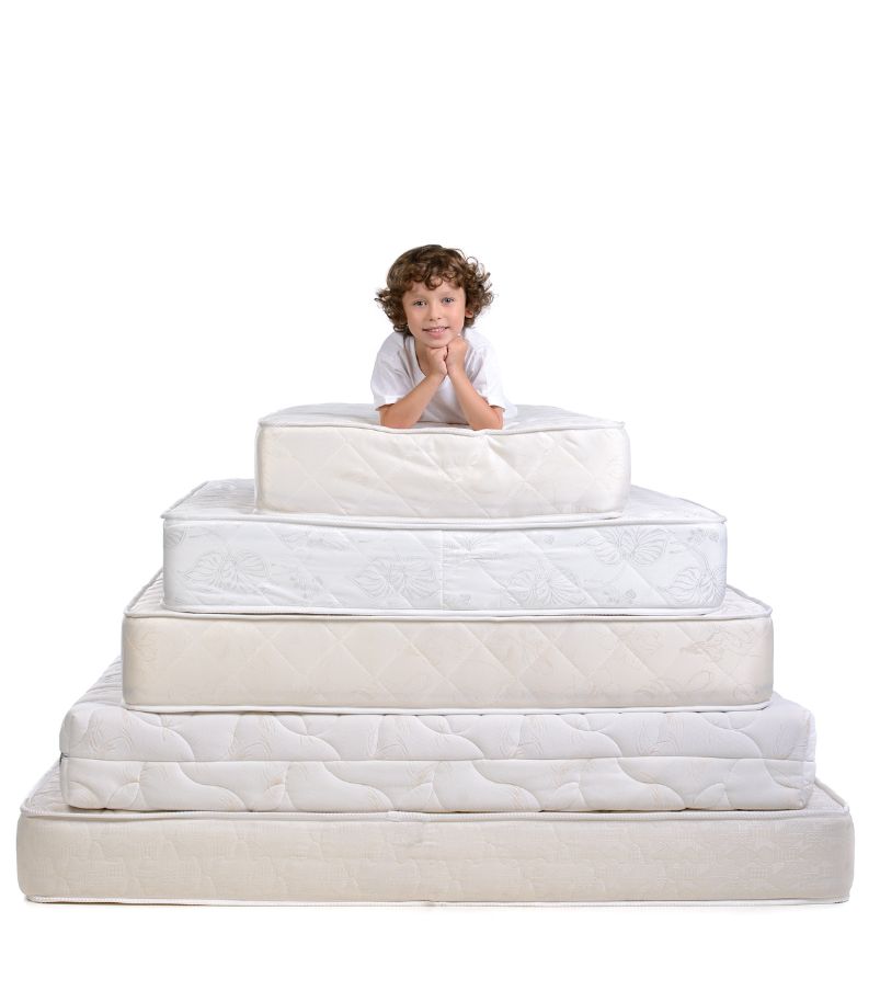 tamanho-e-medidas-de-colchoes Pilha de colchões de diferentes tamanho e medidas de colchões, com uma criança deitada no topo para ilustrar a escala e variedade de opções.