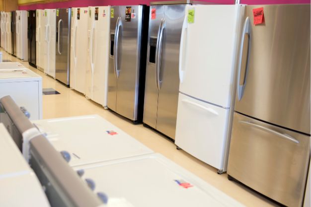 Fileira de geladeiras expostas em loja, com modelos em aço inox e branco. Eletrodomésticos de duas portas alinhados lado a lado em showroom iluminado. Geladeiras novas à venda, com etiquetas de preço visíveis na parte frontal.