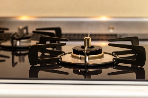 Boca de fogão a gás vista de perto, com queimadores metálicos circulares e suportes de panela pretos sobre mesa de vidro. O queimador central está desmontado, com o bico e a tampa visíveis, indicando manutenção ou limpeza. Superfície limpa e bem iluminada, sem chama acesa.