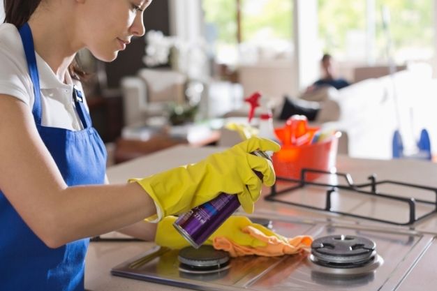Pessoa usando luvas amarelas limpa um fogão a gás com produto em spray e pano de limpeza. As bocas do fogão metálicas estão visíveis sobre a mesa, sem chamas acesas. A cena ocorre em uma cozinha residencial bem iluminada.