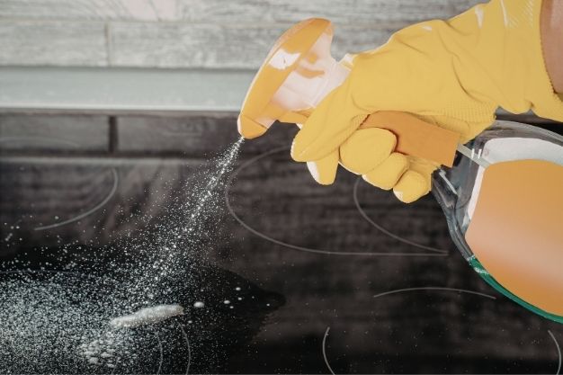 Mão usando luva amarela borrifa produto de limpeza sobre fogão com superfície de vidro preto. O líquido é aplicado diretamente na área engordurada, com partículas visíveis se espalhando sobre a mesa do fogão. A cena mostra o início do processo de remoção de gordura.