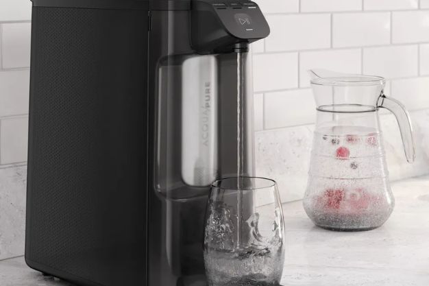 Purificador de água preto sobre bancada servindo água em copo de vidro ao lado de jarra