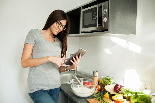 Mulher utilizando tablet na cozinha ao lado de micro-ondas embutido e ingredientes sobre a bancada